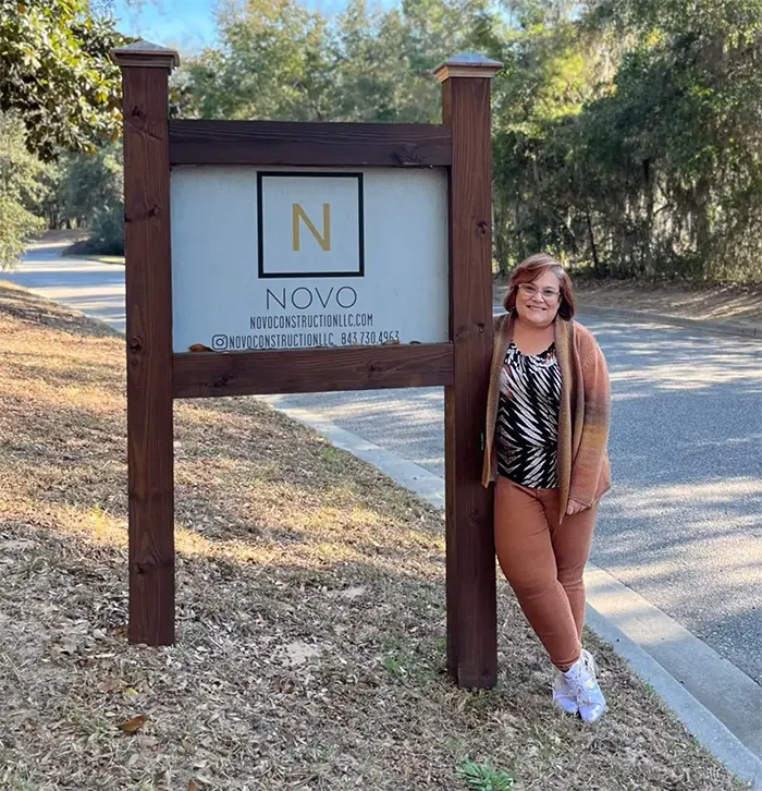 A person stands smiling next to a wooden sign for Novo Construction LLC on a roadside. The sign features a large "N" logo. Trees and greenery are visible in the background, and the person is wearing a patterned shirt and orange pants.