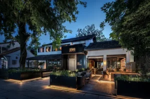 A modern restaurant at dusk features a sleek, minimalist design with large glass windows and an outdoor seating area. Tall trees frame the building, and soft lighting illuminates the pathway and greenery, creating a cozy and inviting atmosphere.