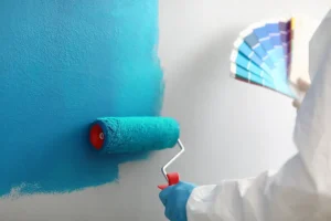 A person wearing gloves and white clothing is painting a wall with blue paint using a roller. They are also holding a color fan deck with various shades of blue in their other hand. The partially painted wall shows a transition from white to blue.